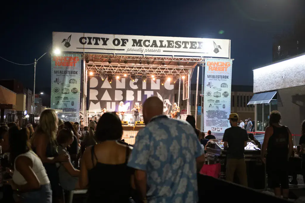 Nighttime outdoor concert stage with crowd gathered in front, illuminated by stage lights and venue signage.
