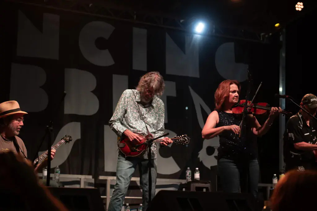 A band performs on stage with guitar and violin players under bright stage lights and large signage backdrop.