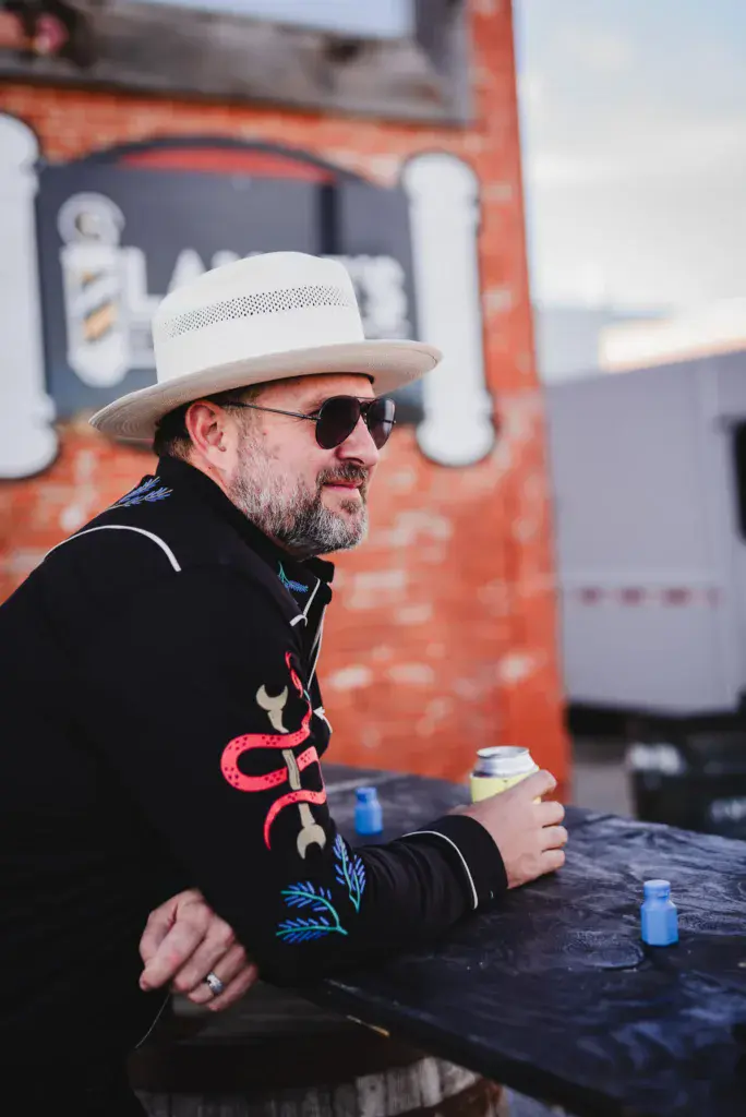 A man in a white cowboy hat and dark embroidered jacket sits at a table holding a beverage at an outdoor festival.