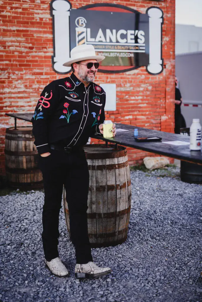 A man in a white cowboy hat and ornate black embroidered jacket holds a beverage near a wooden barrel.