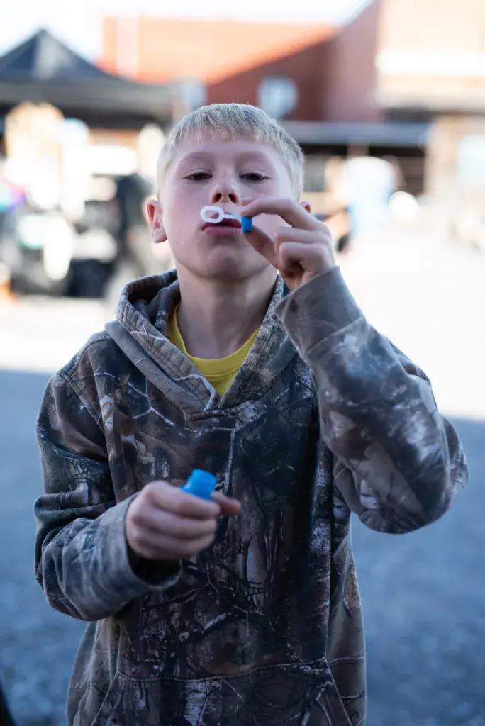 A young child blows a blue whistle while wearing a tie-dye jacket at an outdoor festival.