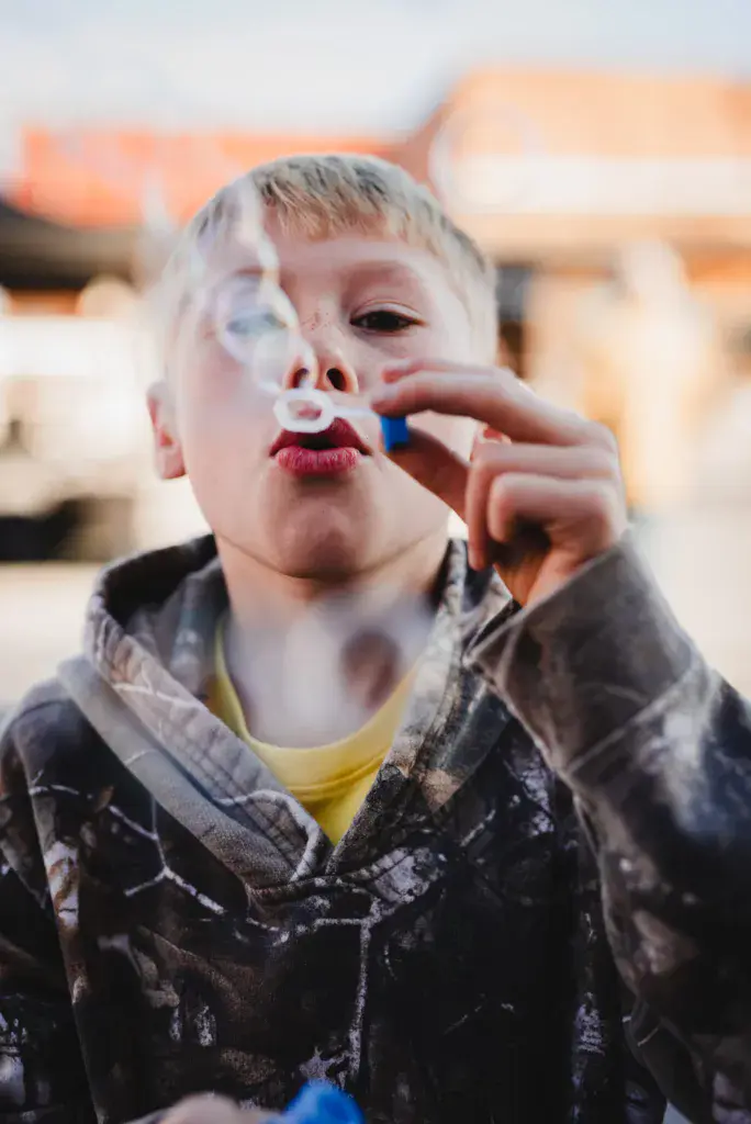 A young child blows bubbles at an outdoor festival venue with colorful structures in the soft-focus background.