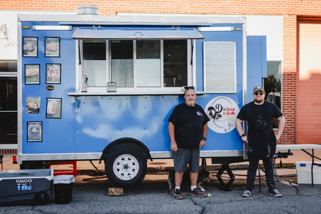 Two people stand in front of a blue food truck vendor at an outdoor festival.