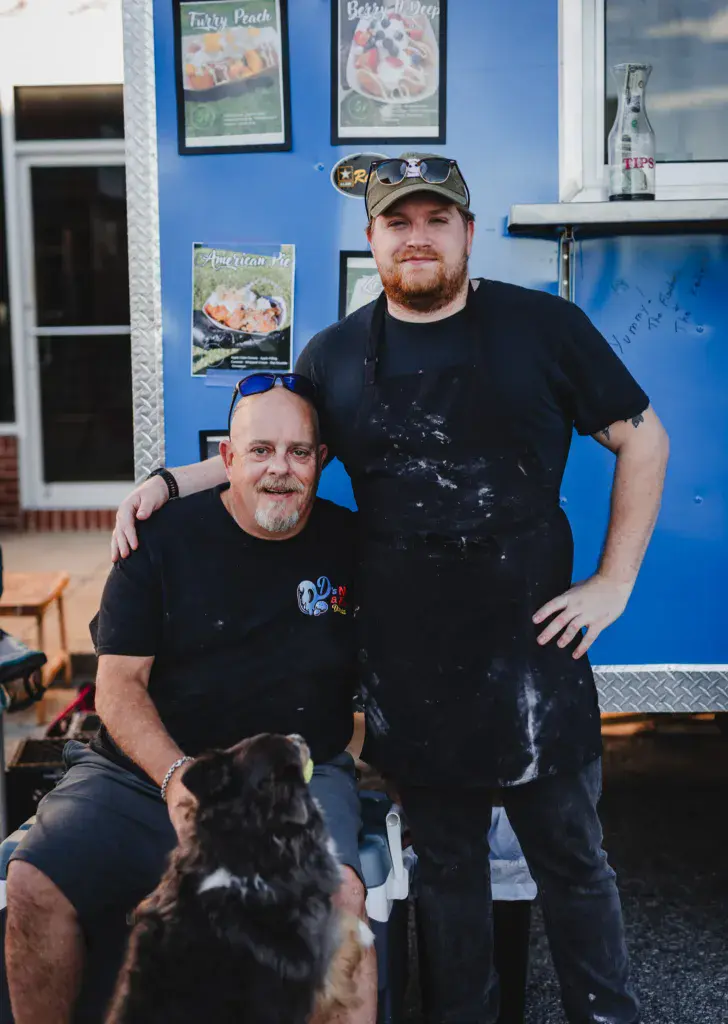 Two people posing together inside a blue food vendor trailer with menu boards on the walls.