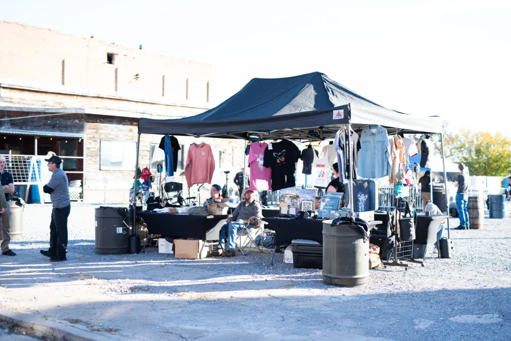 A merchandise vendor tent with clothing displays and staff under a blue canopy at an outdoor festival.