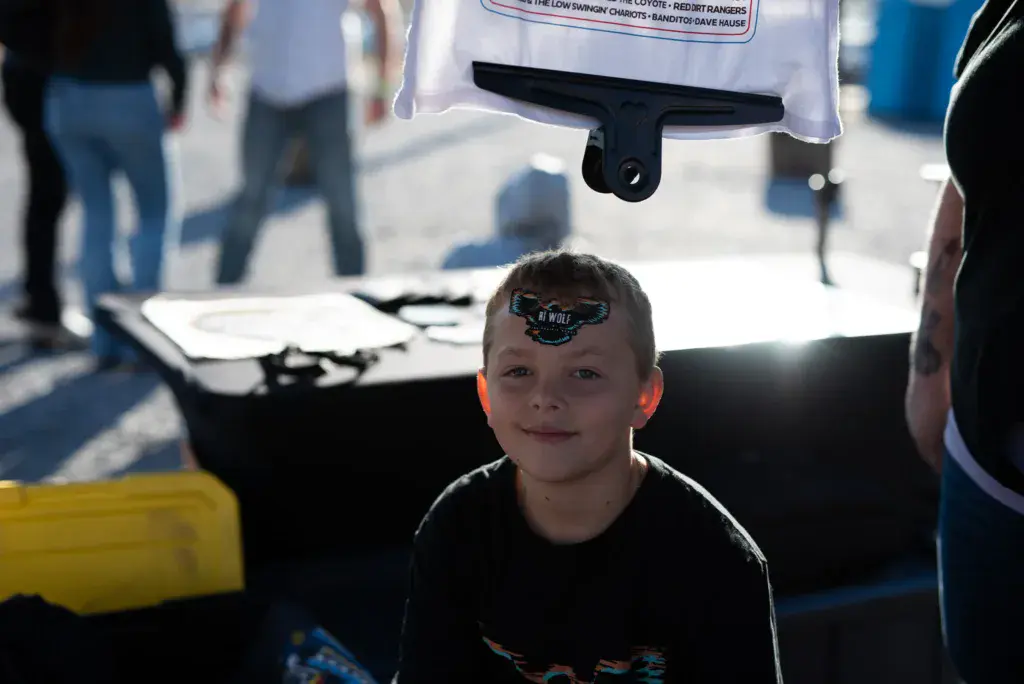 A young child with a festival sticker on their forehead sits under a vendor tent at an outdoor event.