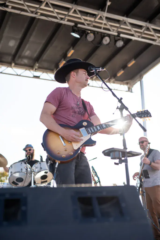 A guitarist in a purple shirt and black cowboy hat sings into a microphone while performing on an outdoor stage.