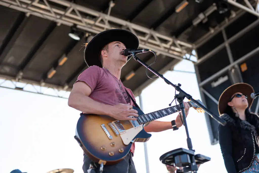A performer in a purple shirt and black cowboy hat plays an electric guitar while singing into a microphone on an outdoor festival stage.