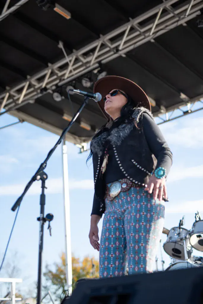A performer sings into a microphone on an outdoor stage, wearing a brown hat, black studded vest, and patterned skirt.