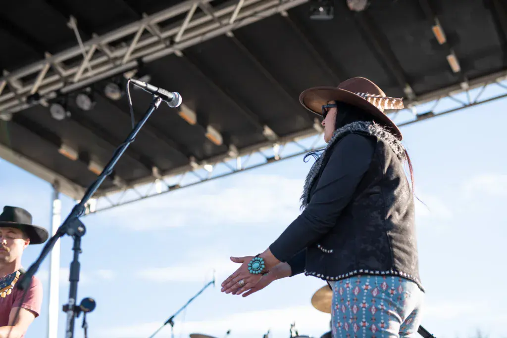 A performer wearing a cowboy hat and ornate vest sings into a microphone on stage under a metal canopy.