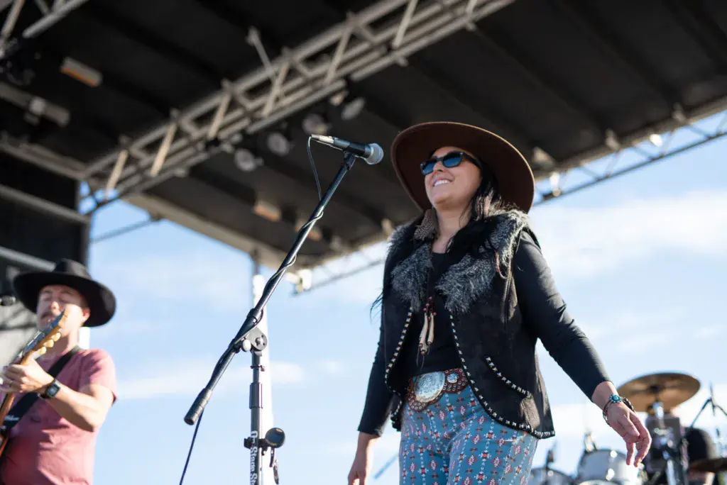 A performer in a brown hat and black jacket sings at a microphone on an outdoor festival stage.