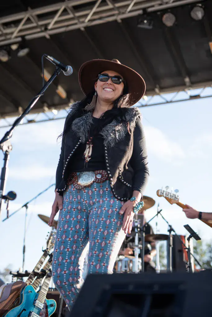 A performer on stage wearing a brown cowboy hat, black studded jacket, and patterned blue pants plays guitar at the festival.