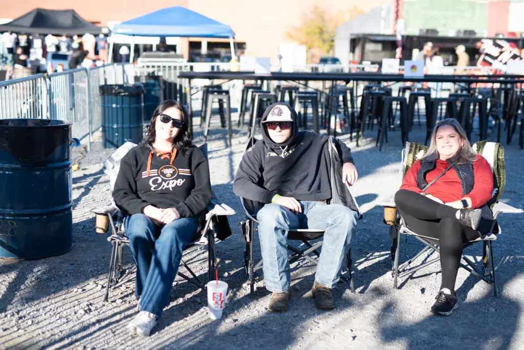 Three attendees sit in lawn chairs in the festival grounds on a sunny day.