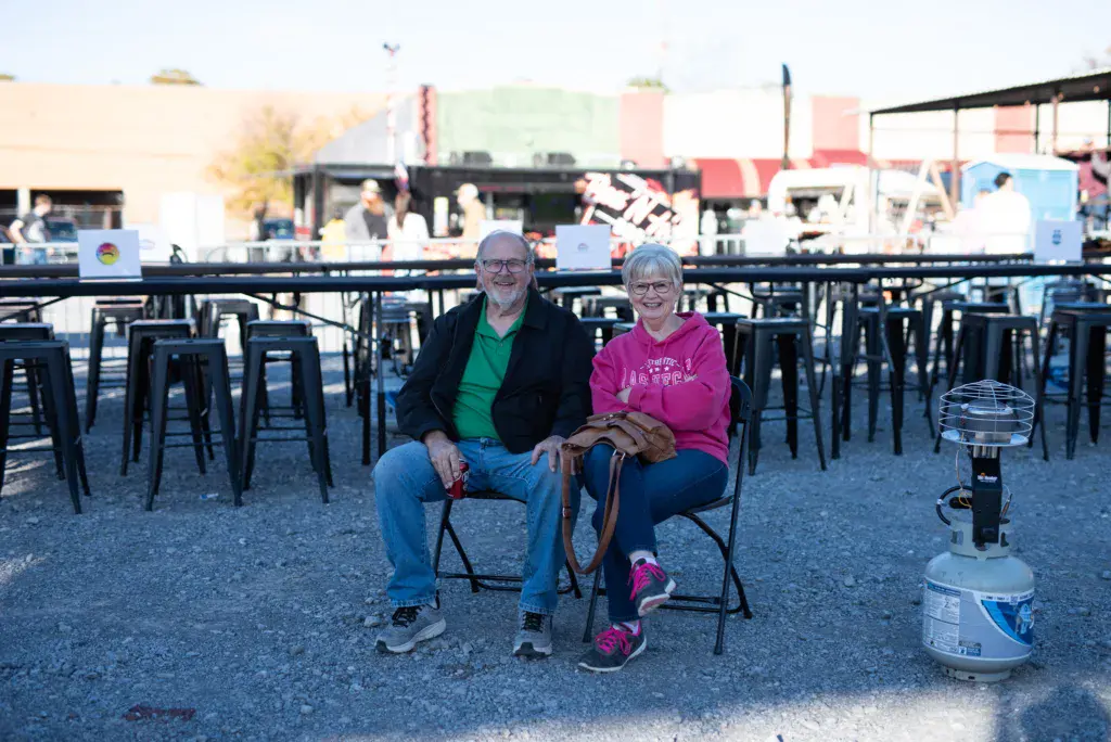 Two people sit together at an outdoor festival venue with festival infrastructure and vendor booths visible in the background.