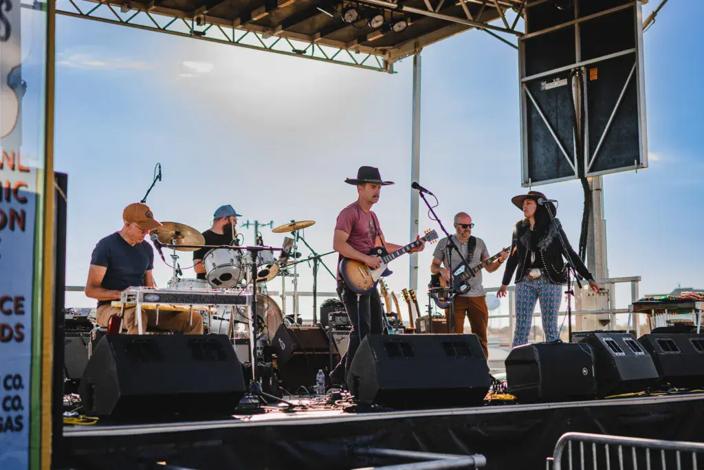 A five-member band performs on an outdoor stage under a metal canopy with blue sky in the background.