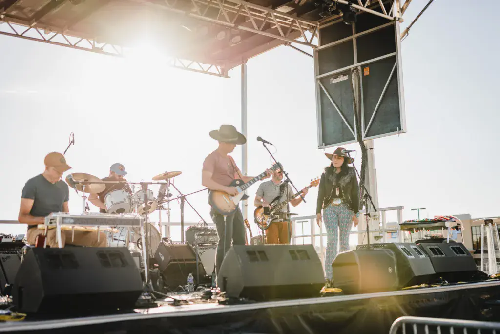 A five-piece band performs on an outdoor stage beneath a metal canopy structure during daytime.