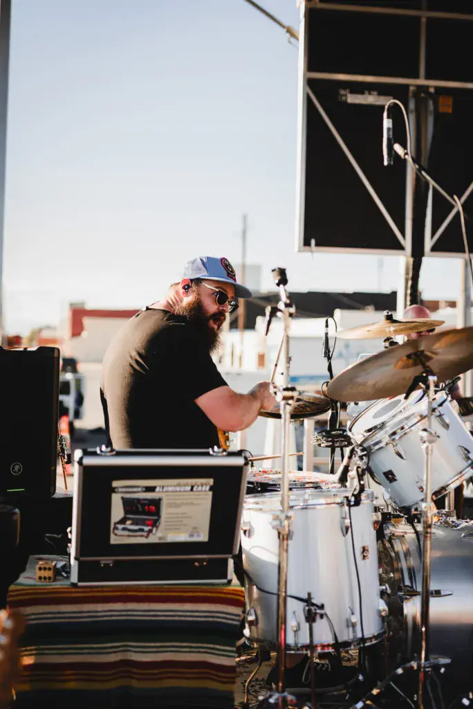 A drummer performing at an outdoor music festival stage, playing a white drum kit in bright sunlight.