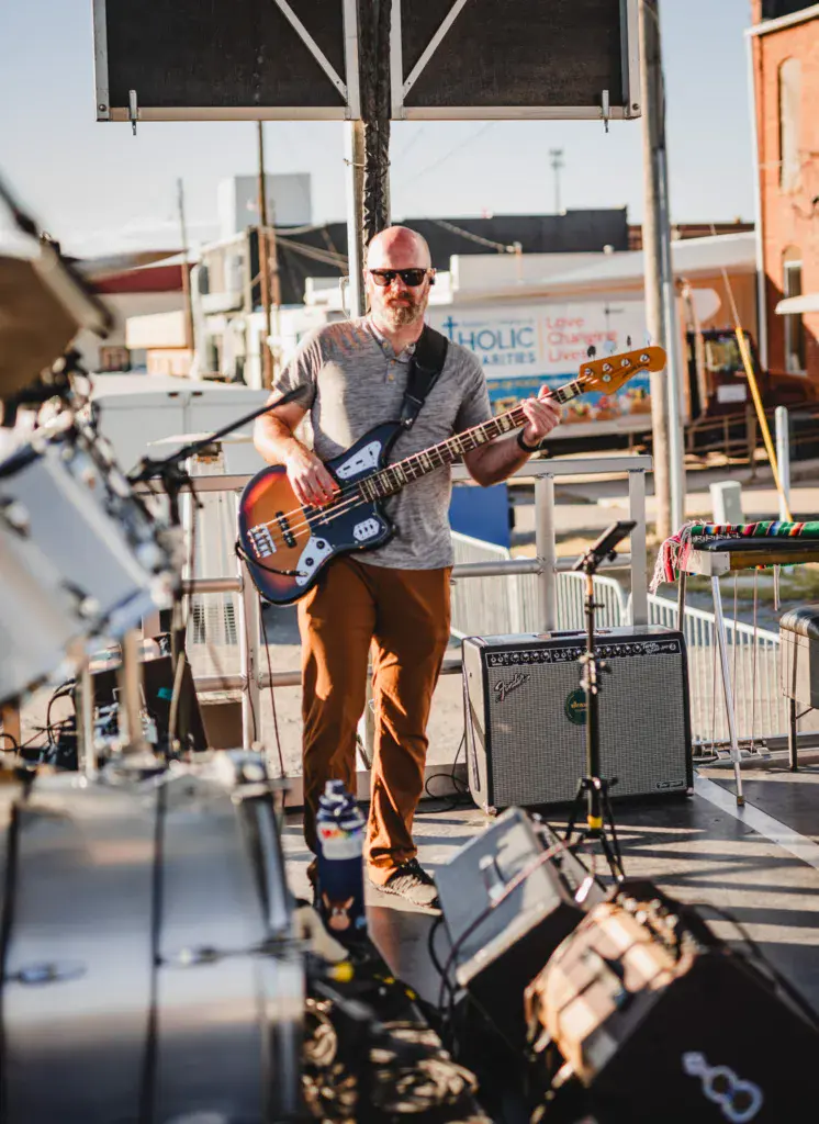A bass guitarist performs on an outdoor stage at a music festival under bright sunlight.