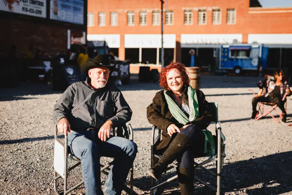 Two people seated in chairs at an outdoor festival venue with orange and blue buildings in the background.