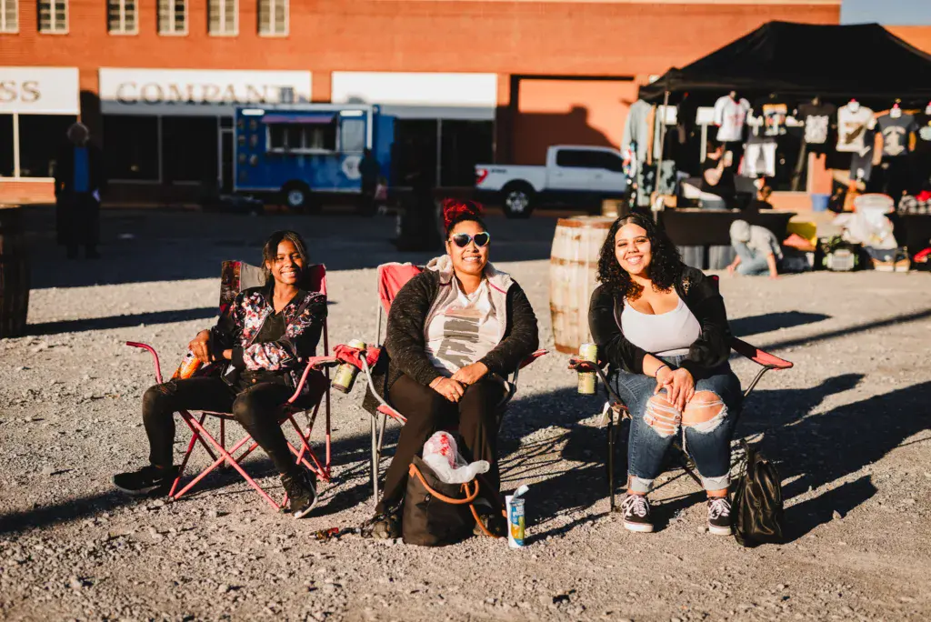 Three people sitting in camping chairs at an outdoor festival venue with food trucks and vendor tents in the background.