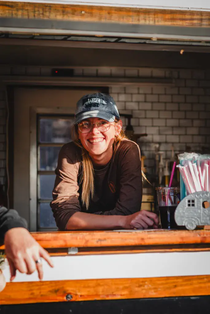 A smiling person wearing a cap and glasses serves drinks from a festival beverage stand.