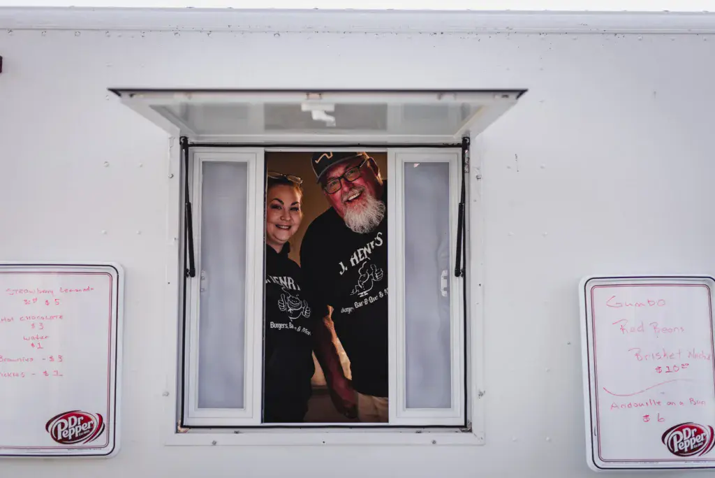 Two people smile from a food vendor window displaying menu boards on either side.