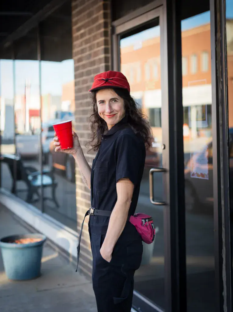 A person in a red cap and dark outfit holds a red cup outside a brick building.
