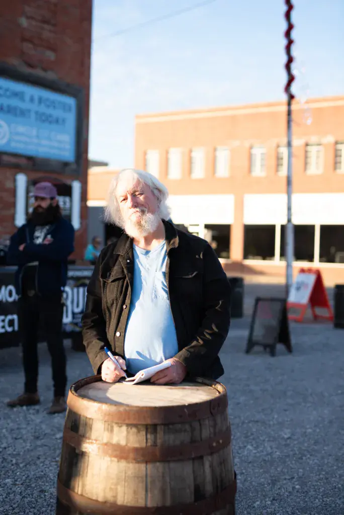 A person with white hair and beard stands on a wooden barrel in a town square during daytime.