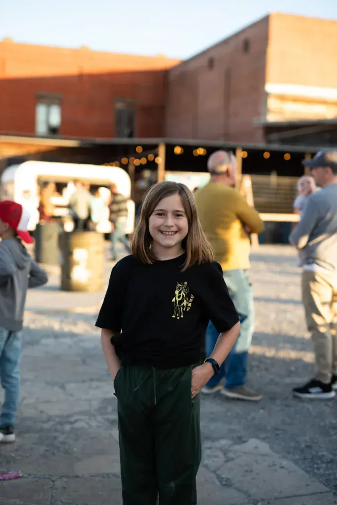 Young girl smiling at camera in festival grounds with food vendors and venue buildings in background