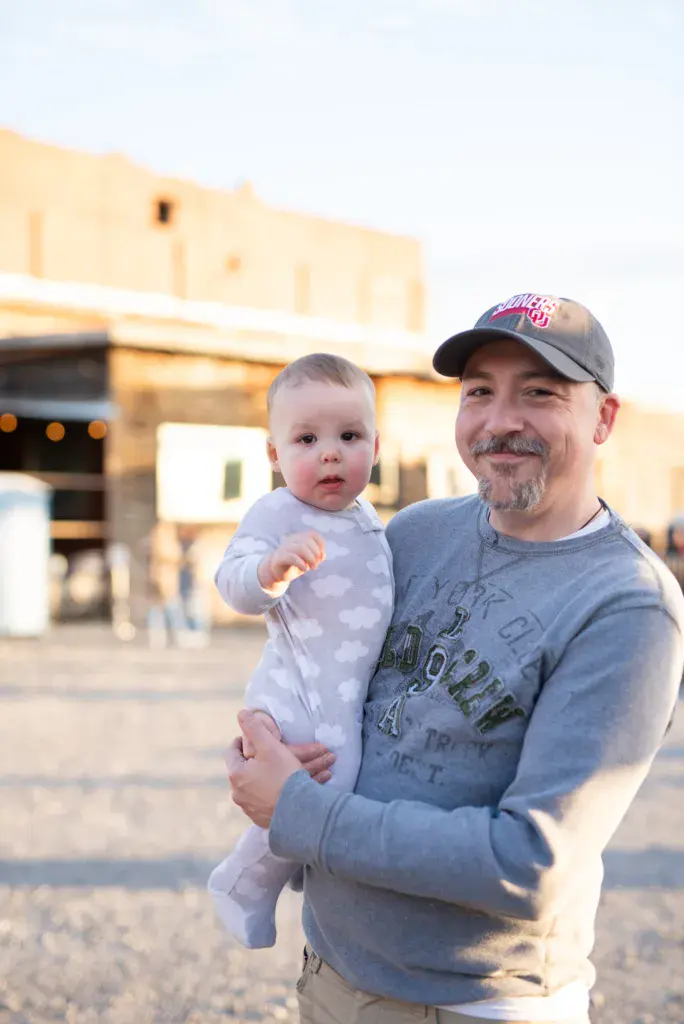An adult holding a young child at an outdoor festival venue with desert-style buildings in the background.