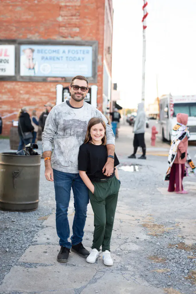 A man and child stand together on a street at the festival with brick buildings and other attendees visible in the background.