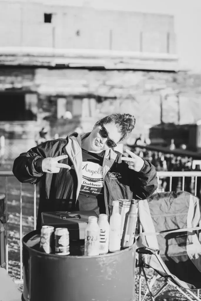 A person wearing sunglasses and a jacket poses playfully at a festival venue with food and drinks nearby.