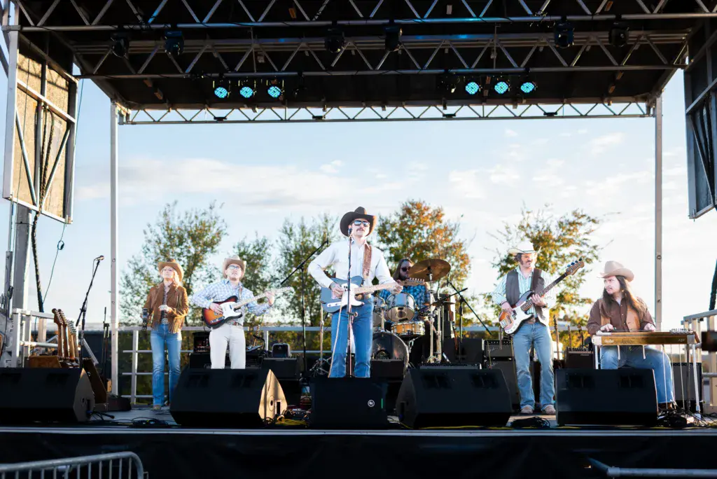 A five-piece country band performs on an outdoor stage under bright lights with autumn trees in the background.