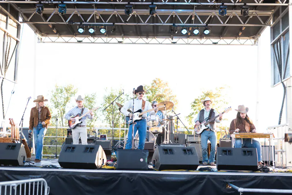 A six-piece band performs on an outdoor stage with guitars, drums, and keyboards under bright stage lighting.