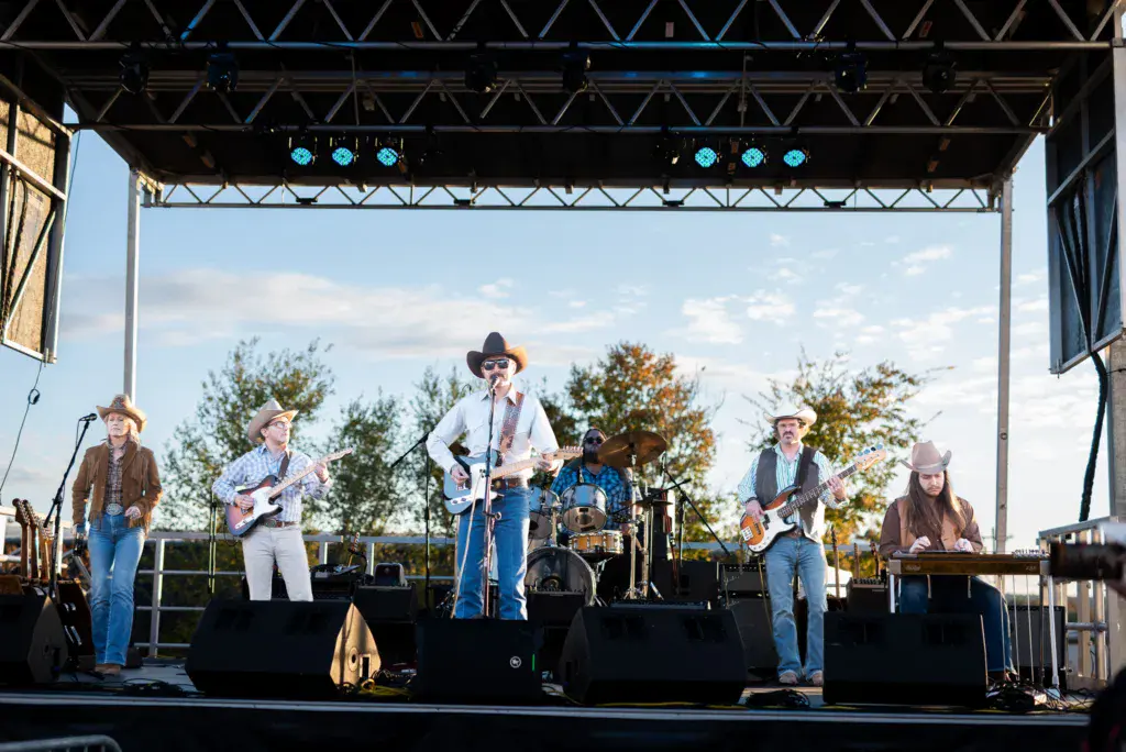 A six-piece band performs on an outdoor stage under a metal truss roof with blue stage lights.