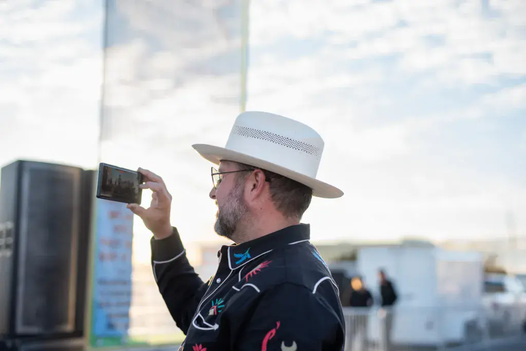 A man in a cowboy hat and decorative jacket takes a photo with his phone at an outdoor festival.