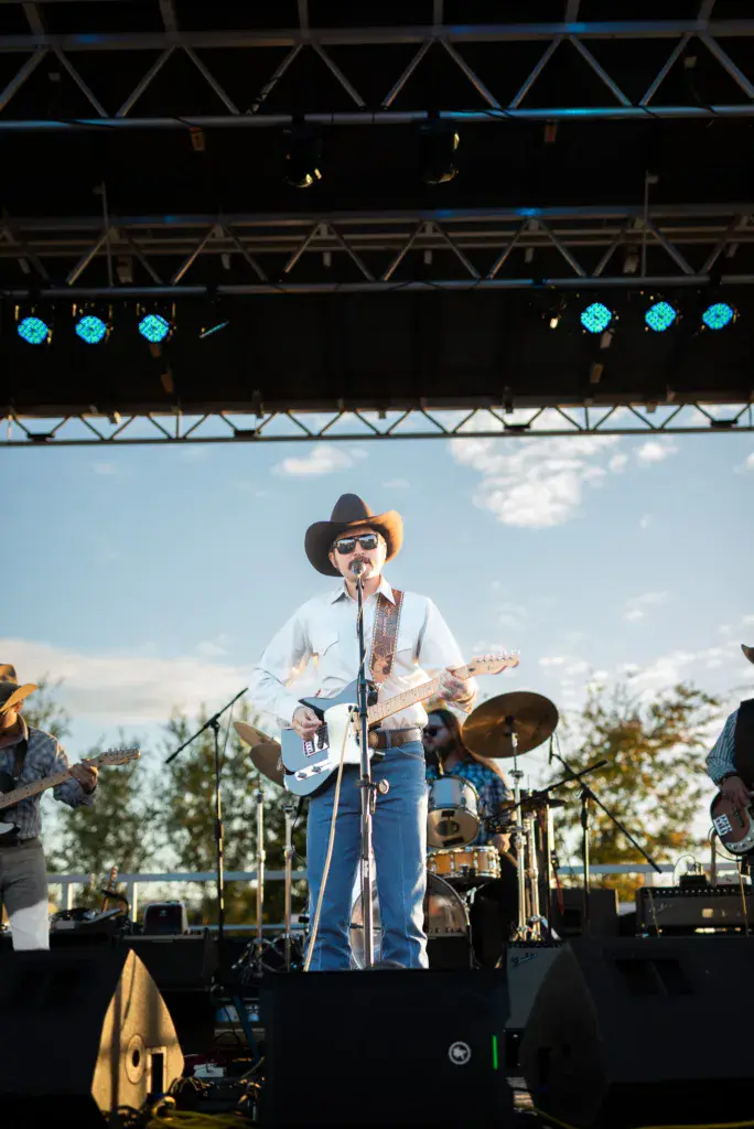 A performer in a cowboy hat and sunglasses plays electric guitar on stage under professional lighting rigs.