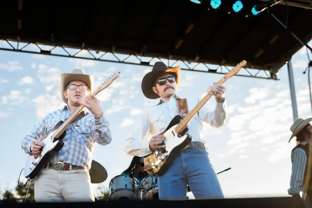 Two guitarists perform on stage under a covered structure with blue lighting and daytime sky visible behind them.