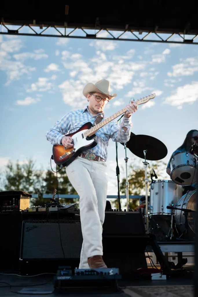 A guitarist in a cowboy hat and glasses performs on an outdoor stage under blue sky with clouds.