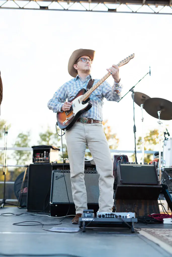 A guitarist wearing a cowboy hat and glasses performs on an outdoor stage at a music festival.