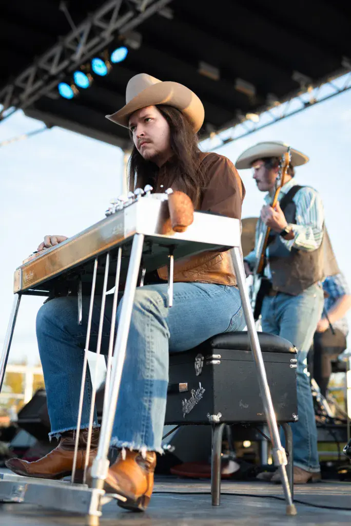 A performer wearing a cream-colored cowboy hat plays pedal steel guitar on stage during the festival.