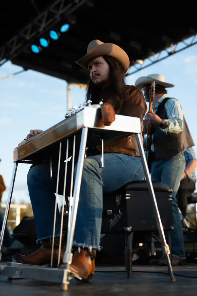 A performer wearing a tan cowboy hat and brown vest plays guitar on stage during the festival.