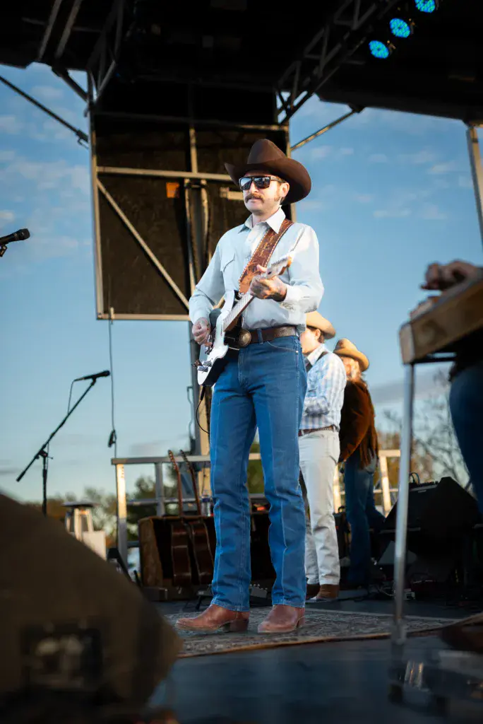 Musician in cowboy hat and sunglasses playing guitar on stage under bright blue sky