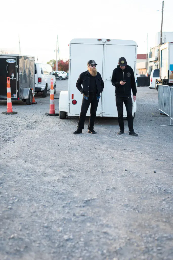 Two people in black clothing stand in front of a white trailer at a festival venue with orange traffic cones visible.