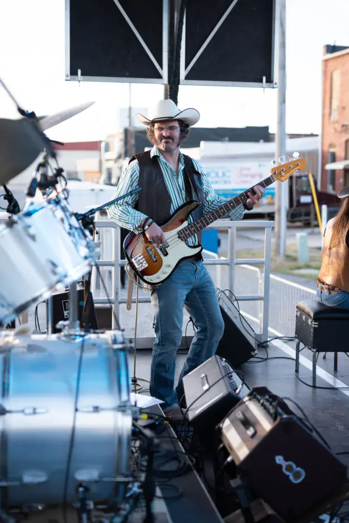 A bass guitarist in a cowboy hat and striped shirt performs on an outdoor stage at the festival.