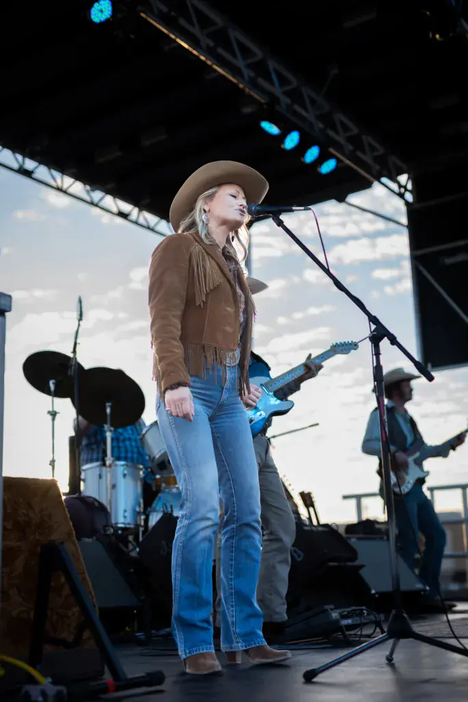A performer wearing a cowboy hat and fringe jacket sings at a microphone on stage with band members and instruments visible behind her.