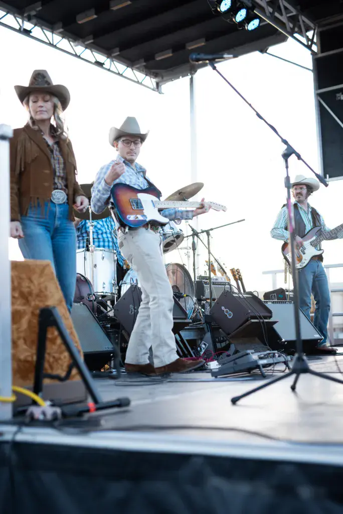 Three musicians in cowboy hats perform on an outdoor festival stage with guitars and drums under stage lighting.