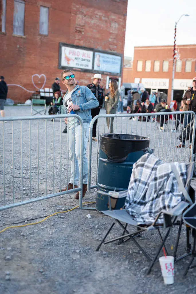 A person in a denim shirt and sunglasses stands near a fire barrel at an outdoor festival venue with brick buildings and crowds visible.