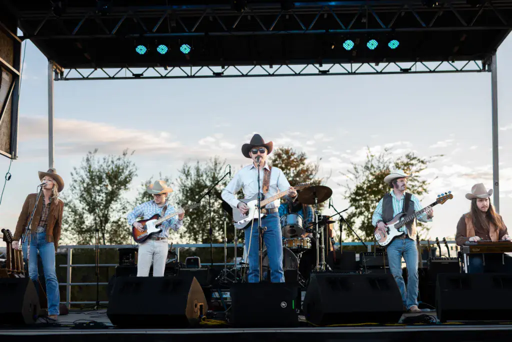 A five-piece country band performs on an outdoor stage with blue lights and metal truss rigging.