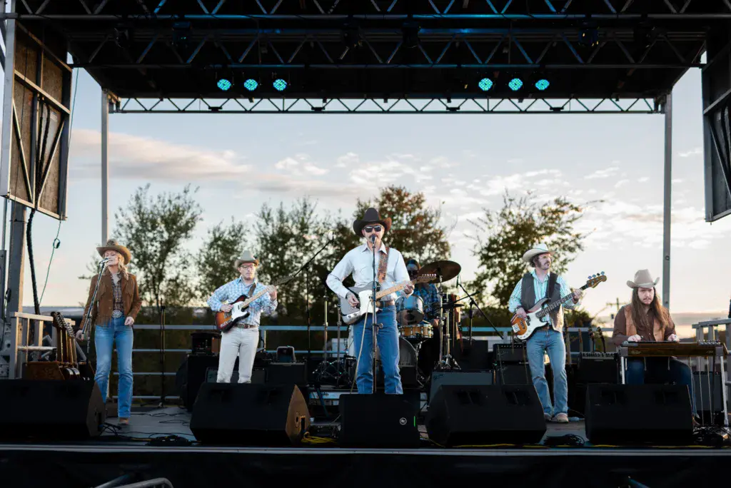 A five-piece band performs on an outdoor stage with blue lighting rigs and scenic backdrop.